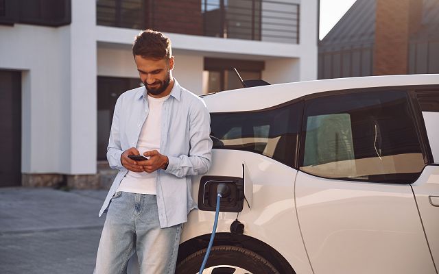 Fertighaus Einfamilienhaus Ladesituation Elektroauto, Person steht am Fahrzeug und bedient Smartphone während Ladevorgang im Außenbereich