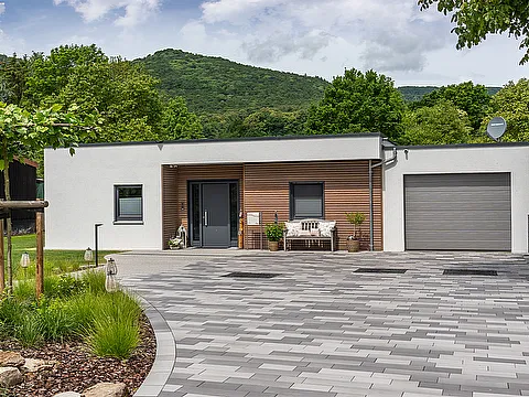 Fertighaus Bungalow mit Flachdach, weißer Fassade, Holzakzent, Garage, gepflasterter Einfahrt, grüner Hanglage