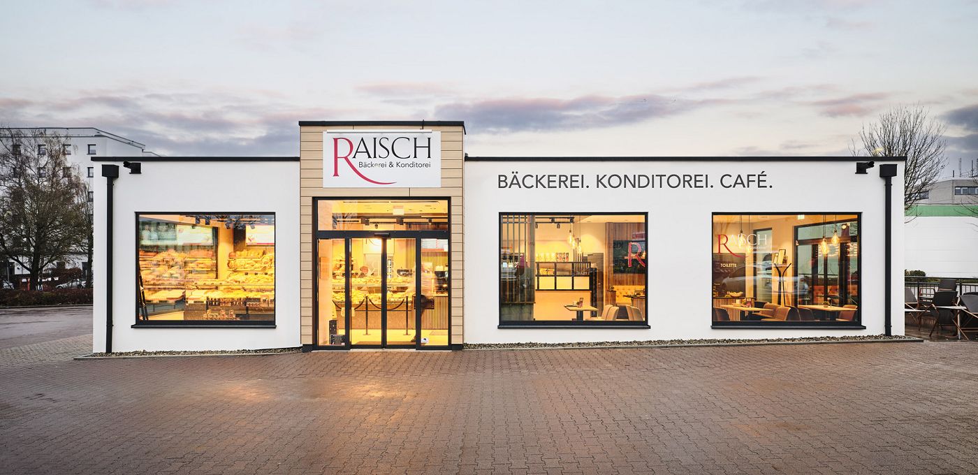 Fertighaus Gewerbebau Bakery Cube Bäckerei mit Putzfassade, großen Schaufenstern und zentralem Eingangsbereich bei Dämmerung